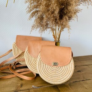 May include: Three round straw bags with leather flap closures and straps. The bags are arranged in a row on a wooden surface.
