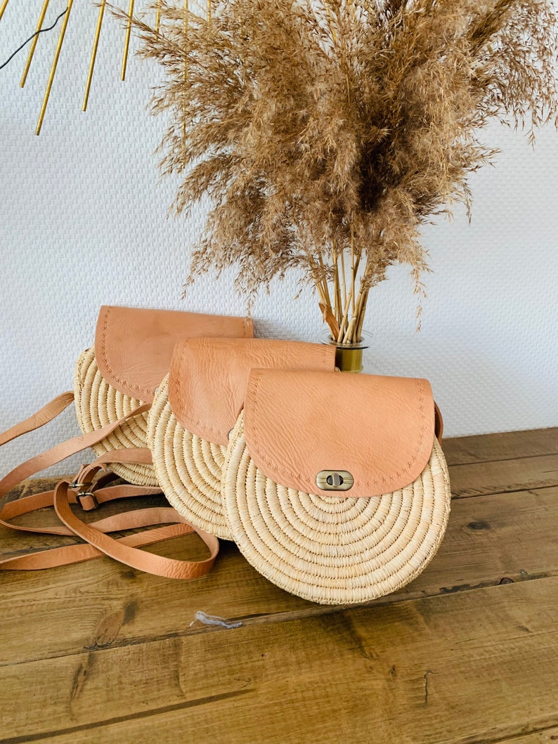 May include: Three round straw bags with leather flap closures and straps. The bags are arranged in a row on a wooden surface.