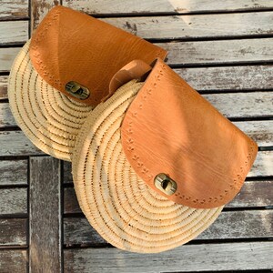 May include: Two round straw bags with brown leather flaps and metal closures. The bags are sitting on a wooden surface.
