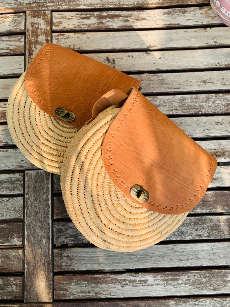 May include: Two round straw bags with brown leather flaps and metal closures. The bags are sitting on a wooden surface.