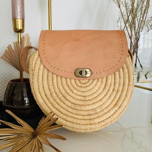 May include: A round straw bag with a tan leather flap and a brass clasp. The bag has a woven design and a strap.