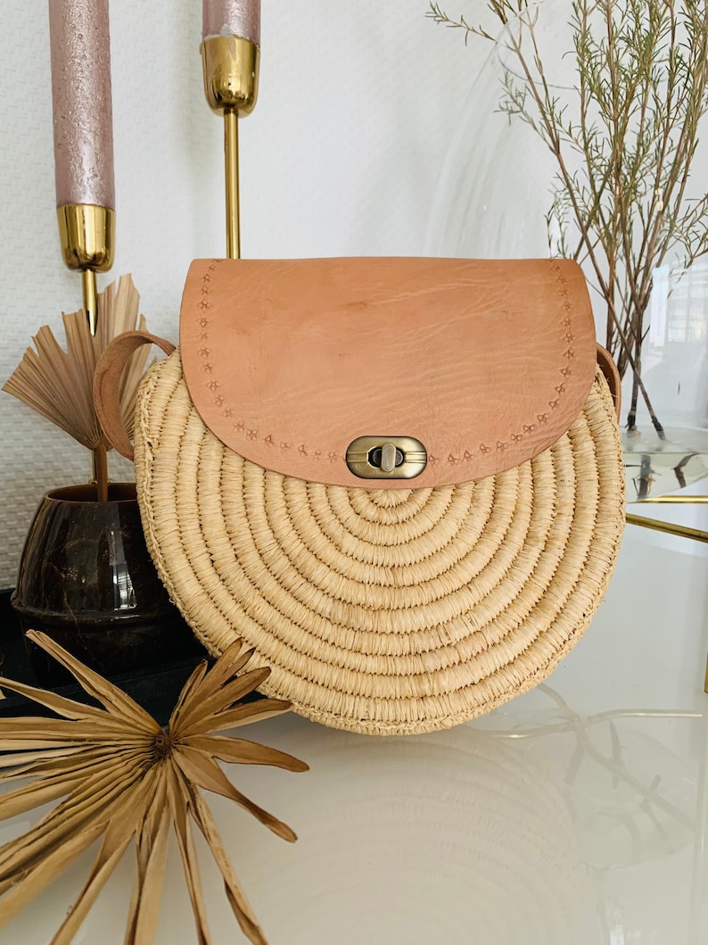 May include: A round straw bag with a tan leather flap and a brass clasp. The bag has a woven design and a strap.