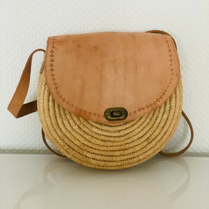 May include: A round straw bag with a brown leather flap and a metal clasp. The bag has a long brown leather strap.