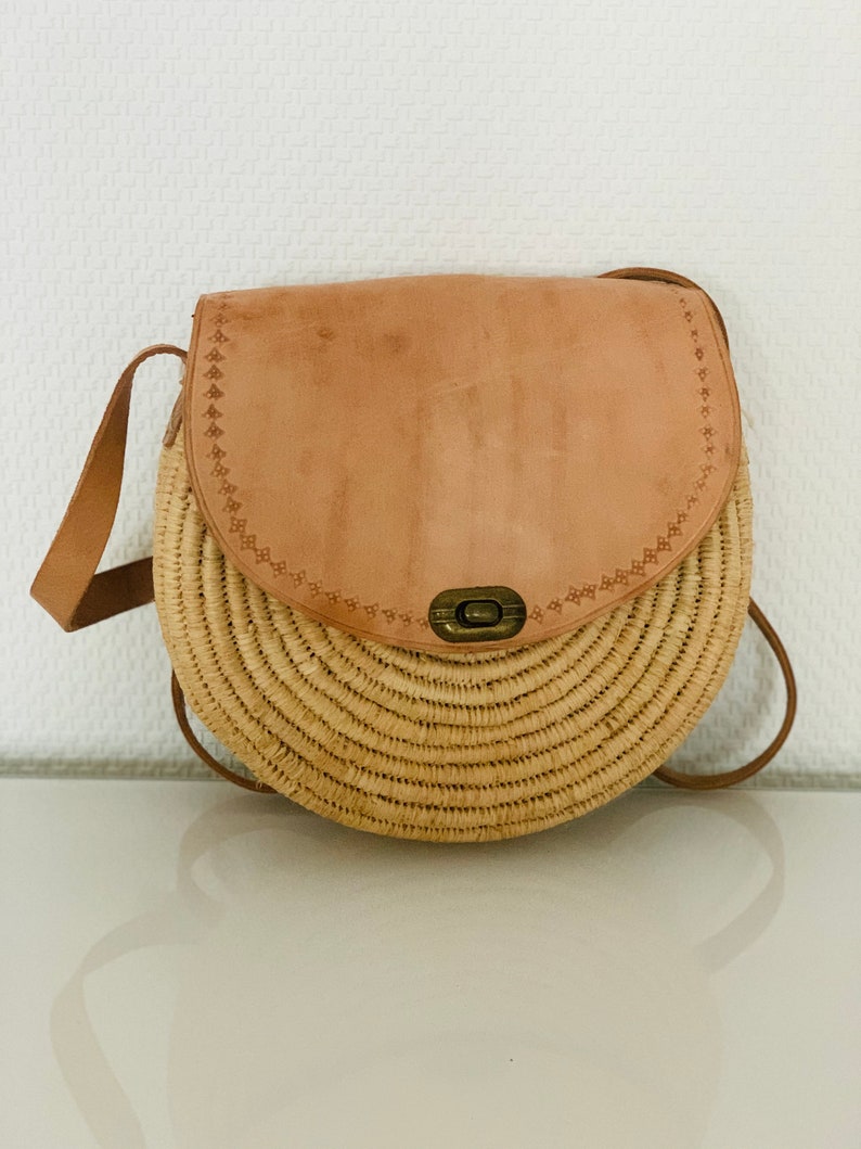 May include: A round straw bag with a brown leather flap and a metal clasp. The bag has a long brown leather strap.