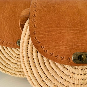 May include: A round straw bag with a brown leather flap and a brass clasp. The leather flap has a decorative stitched pattern.