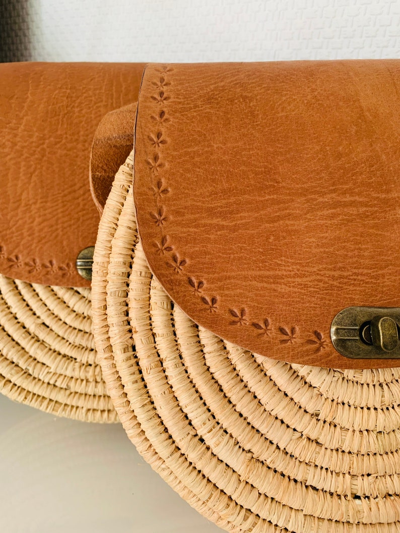 May include: A round straw bag with a brown leather flap and a brass clasp. The leather flap has a decorative stitched pattern.