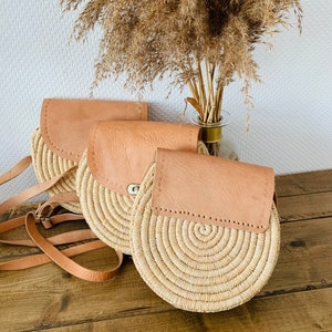 May include: Three round straw bags with leather flap closures. The bags are beige and brown and have a woven texture. The bags are arranged in a row on a wooden surface.