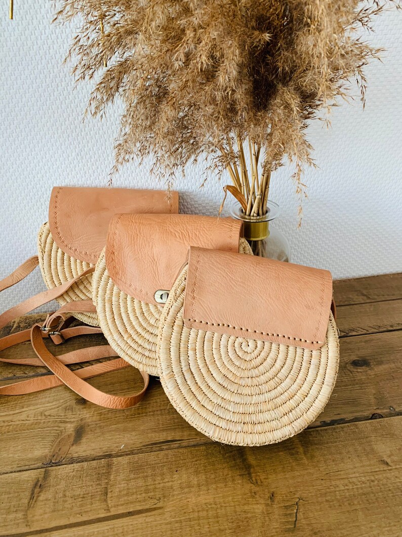 May include: Three round straw bags with leather flap closures. The bags are beige and brown and have a woven texture. The bags are arranged in a row on a wooden surface.