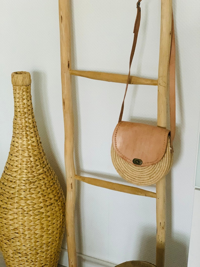 May include: A light brown leather and woven straw crossbody bag hanging on a wooden ladder.