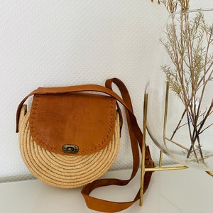 May include: A round straw bag with a brown leather flap and strap. The bag has a brass clasp and is perfect for carrying everyday essentials.