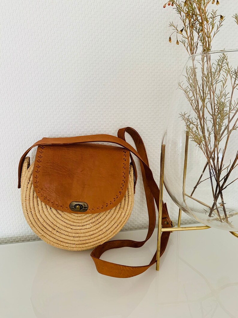 May include: A round straw bag with a brown leather flap and strap. The bag has a brass clasp and is perfect for carrying everyday essentials.