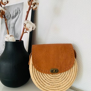 May include: A round straw bag with a brown leather flap and a brass clasp. The bag has a woven design and is perfect for carrying everyday essentials.