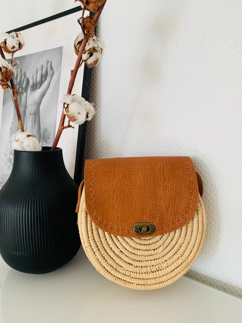 May include: A round straw bag with a brown leather flap and a brass clasp. The bag has a woven design and is perfect for carrying everyday essentials.