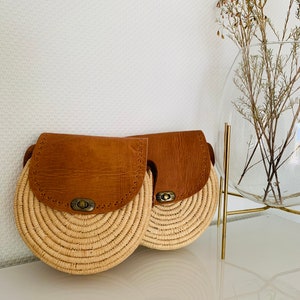 May include: Two round straw bags with brown leather flaps and a brass buckle closure. The bags are sitting on a white surface.