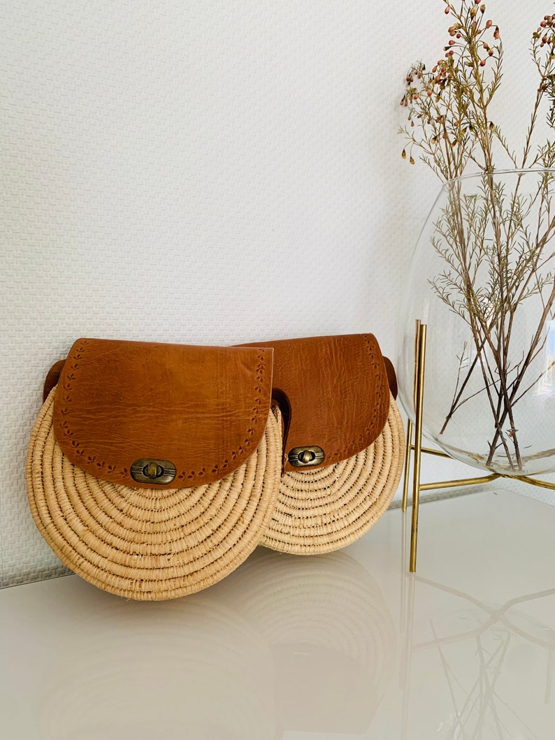 May include: Two round straw bags with brown leather flaps and a brass buckle closure. The bags are sitting on a white surface.