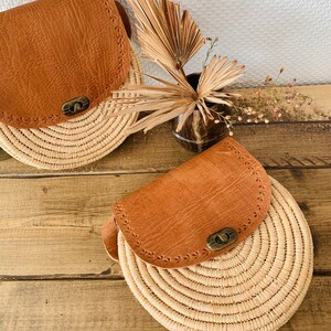 May include: Two round straw bags with brown leather flaps and a brass closure. The bags are on a wooden surface.