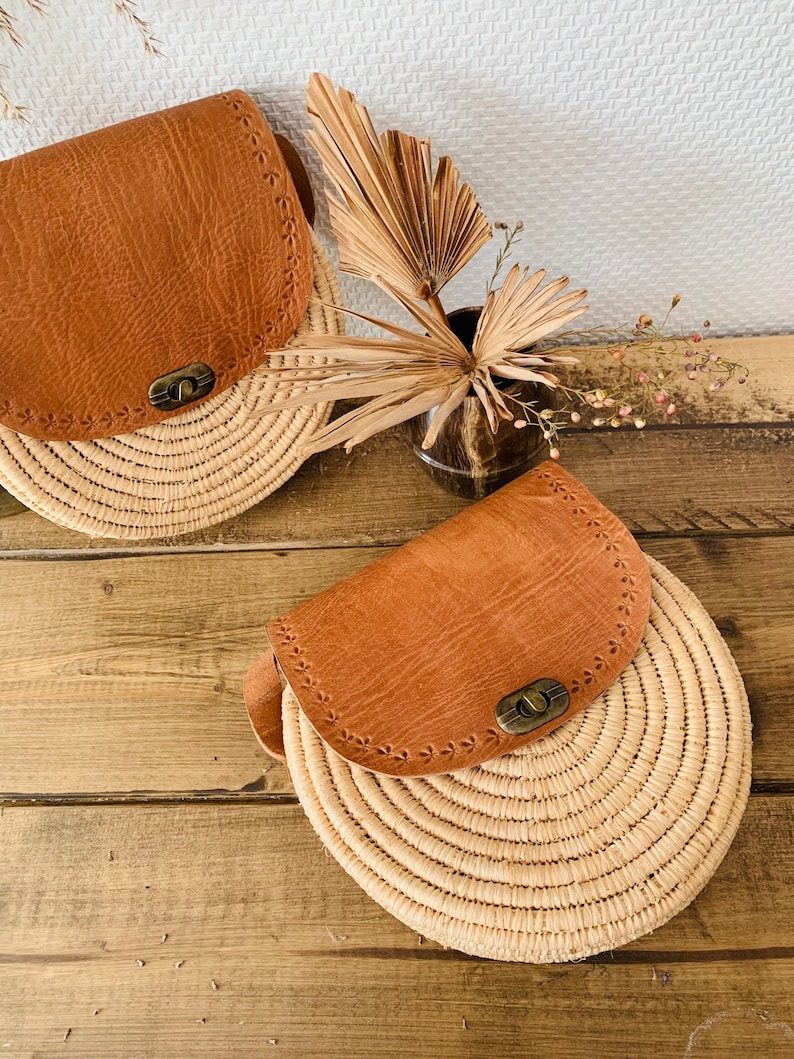 May include: Two round straw bags with brown leather flaps and a brass closure. The bags are on a wooden surface.