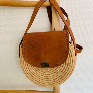 May include: A round straw bag with a brown leather flap and a metal clasp. The bag has a long brown leather strap.