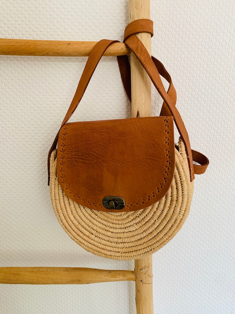 May include: A round straw bag with a brown leather flap and a metal clasp. The bag has a long brown leather strap.