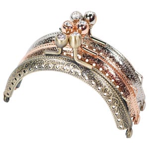May include: An assortment of decorative purse clasps in various metallic finishes, including antique brass, rose gold, and silver. Each clasp features an ornate, openwork design and is embellished with small, clear rhinestones. The clasps are arranged in a layered display.