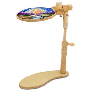 May include: A wooden embroidery stand with a landscape design in an oval hoop. The stand has a curved base, adjustable height, and a wooden knob for securing the hoop. The embroidery features a colorful sunset over mountains.