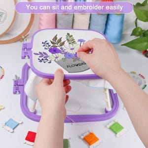 May include: A purple embroidery hoop with a white fabric stretched inside, featuring a floral design with the word "FLOWERS". The image also shows spools of thread in various colors, and the text "You can sit and embroider easily".