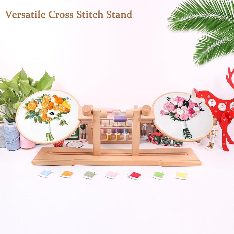 K&ouml;nnte beinhalten: Ein h&ouml;lzerner Kreuzstichst&auml;nder mit zwei Stickrahmen, einer mit einem gelben und orangefarbenen Blumendesign und der andere mit einem rosa und wei&szlig;en Blumendesign. Der St&auml;nder hat ein Regal zum Aufbewahren von Stickgarn.