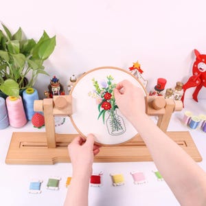 K&ouml;nnte beinhalten: Ein Holzst&auml;nder f&uuml;r einen Stickrahmen mit einem wei&szlig;en Stickrahmen, der ein Blumenmuster zeigt. Der Rahmen wird von den H&auml;nden einer Person gehalten. Auf dem Tisch liegen mehrere Spulen mit Stickgarn.