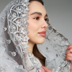 May include: A white sheer head scarf with silver floral embroidery. The scarf is draped over a person's head and shoulders.