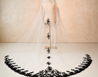 May include: A long, white bridal veil with a delicate black floral lace trim along the bottom edge. The veil is displayed on a mannequin, showcasing the intricate design and flowing fabric. The veil's train extends across the floor.
