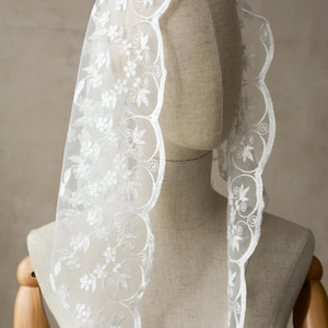 May include: A white, sheer veil with an intricate floral and swirl embroidered design. The veil has a scalloped edge, adding a delicate touch. The veil is draped over a mannequin head.