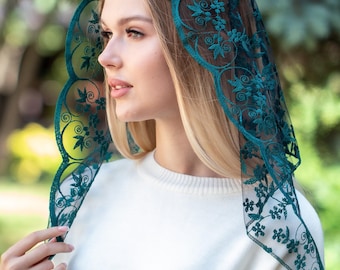 May include: A teal lace head covering with floral patterns. The delicate veil is draped over the head and shoulders, with a scalloped edge. The woman in the image is wearing a white jumper.