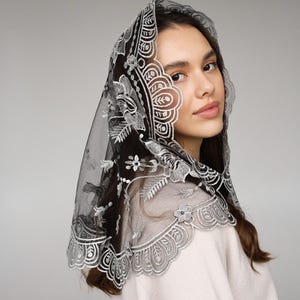 May include: A black veil with intricate white floral and geometric embroidery. The veil is draped over the head and shoulders, showcasing a delicate, lace-like design. The edges are scalloped, adding a touch of elegance.