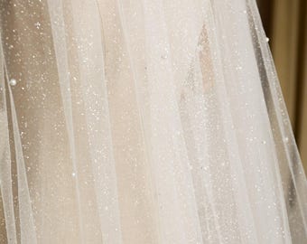 May include: Close-up of a sheer, white bridal veil with a delicate, shimmering glitter effect. The veil is adorned with small, scattered pearls, adding a touch of elegance. The fabric drapes softly, creating a romantic and ethereal look.