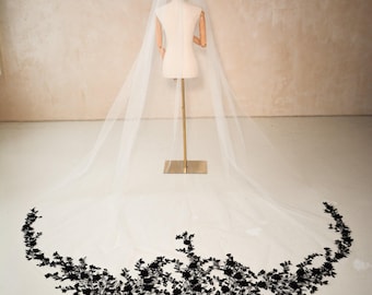 May include: A long, white bridal veil with a delicate black floral lace trim. The veil is displayed on a mannequin, showcasing its length and design. The lace trim is detailed and adds a touch of elegance.
