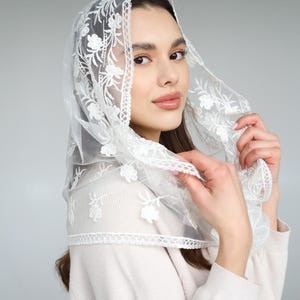 May include: A sheer white veil with delicate floral embroidery. The veil is draped over the head and shoulders, with intricate lace detailing along the edges. The fabric appears soft and lightweight, creating an elegant and ethereal look.