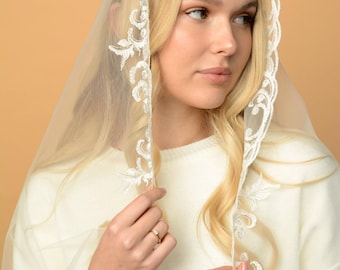 May include: A sheer white veil with delicate floral lace trim. The veil is draped over the head and shoulders, with the lace detailing framing the face. The model is wearing a white sweater.