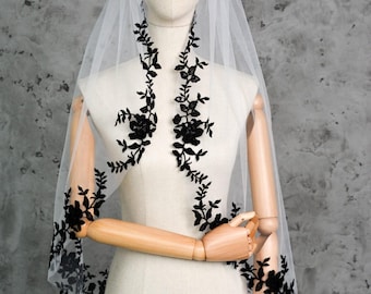 May include: A white bridal veil with black floral lace detailing. The lace is arranged along the edges and down the center, creating a delicate and elegant design. The veil is displayed on a mannequin.