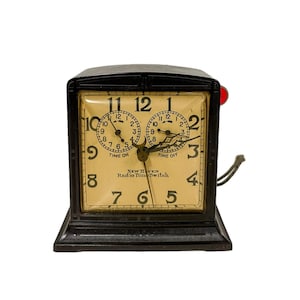 May include: A vintage black New Haven Radio Time Switch clock with a square face. The clock has two dials, one labeled "Time On" and the other "Time Off." The clock face is cream-colored with black numerals and hands. A red button is on the side.