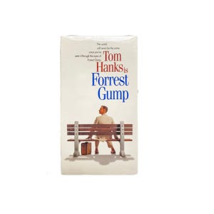 May include: A VHS tape of the movie "Forrest Gump" featuring Tom Hanks. The cover shows a man sitting on a bench with a suitcase, with the film's title in blue and red. The text "The world will never be the same" is also visible.