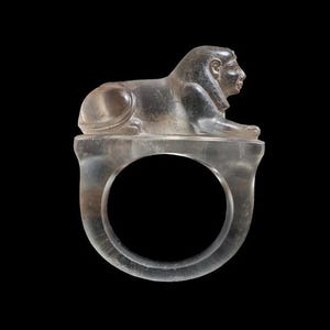May include: A clear ring with a sphinx design. The sphinx, in a reclining position, sits on the ring's band. The ring is made of a transparent material and is set against a black backdrop.