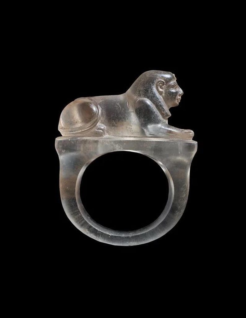 May include: A clear ring with a sphinx design. The sphinx, in a reclining position, sits on the ring's band. The ring is made of a transparent material and is set against a black backdrop.