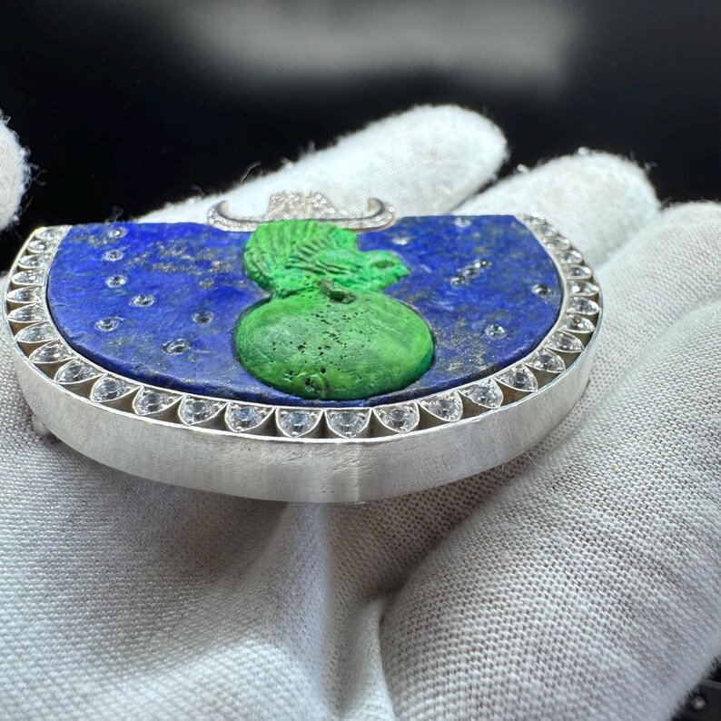 May include: A silver pendant with a blue lapis lazuli base, featuring a green sculpted design. The pendant is edged with small, clear gemstones and has a small, curved silver bail with embedded diamonds.