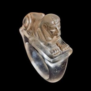 May include: A clear ring with an Egyptian sphinx design. The sphinx has a detailed face and headdress. The ring has a wide band and is set against a black background. The ring appears to be made of a translucent material.