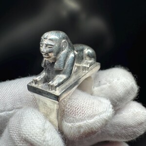 May include: A silver ring with a detailed sphinx design. The sphinx is in a reclining pose on a rectangular base. The ring is made of a shiny, metallic material, and the sphinx has intricate facial features.