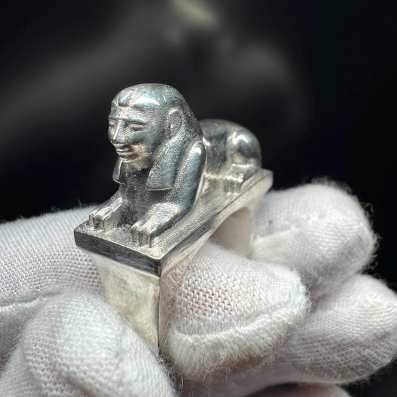 May include: A silver ring with a detailed sphinx design. The sphinx is in a reclining pose on a rectangular base. The ring is made of a shiny, metallic material, and the sphinx has intricate facial features.