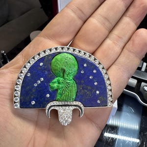 May include: A silver pendant with a blue lapis lazuli background, featuring a green figure. The pendant has a half-circle design with a row of clear stones and a decorative bottom piece, also with clear stones.