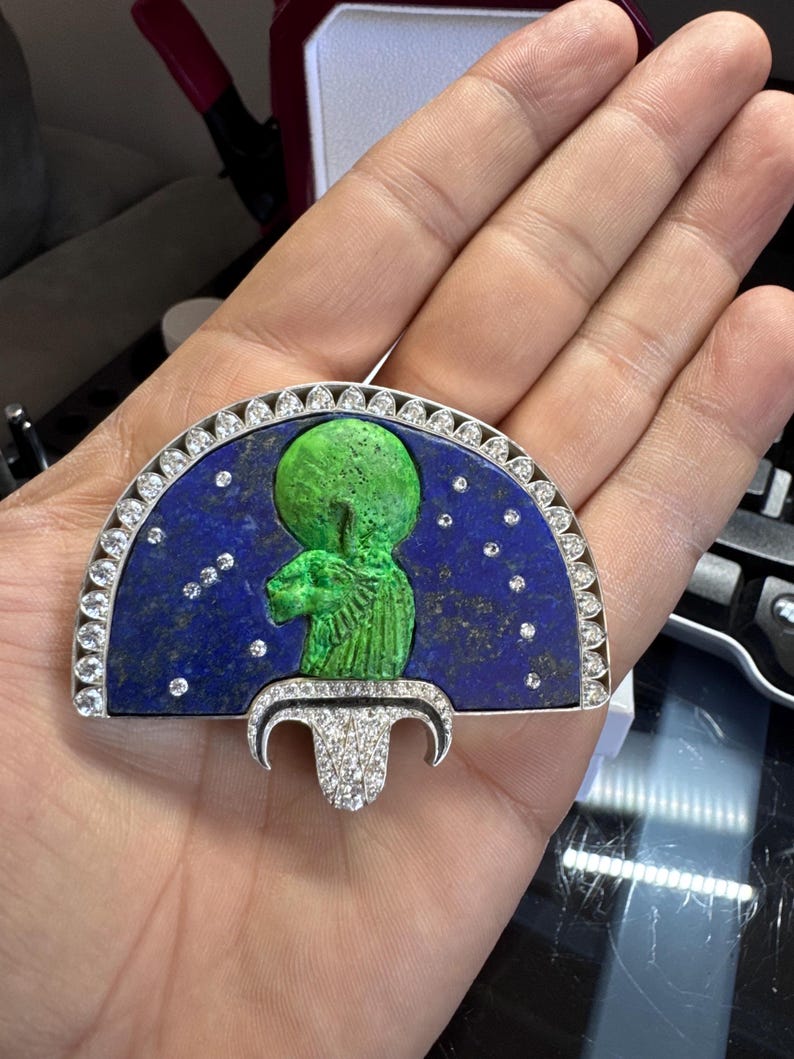 May include: A silver pendant with a blue lapis lazuli background, featuring a green figure. The pendant has a half-circle design with a row of clear stones and a decorative bottom piece, also with clear stones.