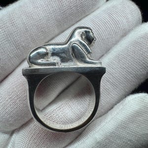 May include: A silver ring with a sphinx design. The sphinx is shown reclining on a rectangular base. The ring's band is a U-shape. The ring has a polished finish, reflecting light. The ring is a piece of jewellery.
