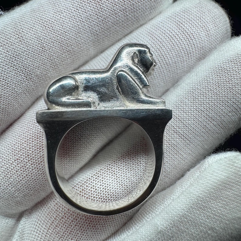 May include: A silver ring with a sphinx design. The sphinx is shown reclining on a rectangular base. The ring's band is a U-shape. The ring has a polished finish, reflecting light. The ring is a piece of jewellery.